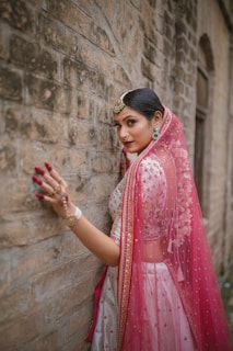 A woman adorned in traditional attire stands gracefully against a textured stone wall. Her outfit features intricate embroidery and vibrant patterns. A sheer, pink veil drapes over her head and shoulders, complemented by elegant jewelry including earrings and a headpiece. Her hand is gently touching the wall, displaying decorative bangles and rings.