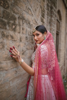A woman adorned in traditional attire stands gracefully against a textured stone wall. Her outfit features intricate embroidery and vibrant patterns. A sheer, pink veil drapes over her head and shoulders, complemented by elegant jewelry including earrings and a headpiece. Her hand is gently touching the wall, displaying decorative bangles and rings.