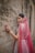 A woman adorned in traditional attire stands gracefully against a textured stone wall. Her outfit features intricate embroidery and vibrant patterns. A sheer, pink veil drapes over her head and shoulders, complemented by elegant jewelry including earrings and a headpiece. Her hand is gently touching the wall, displaying decorative bangles and rings.