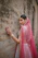 A joyful woman dressed in elegant ethnic wear, standing against a softly lit sandstone wall with subtle Indian textile patterns.