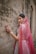 A woman adorned in traditional attire stands gracefully against a textured stone wall. Her outfit features intricate embroidery and vibrant patterns. A sheer, pink veil drapes over her head and shoulders, complemented by elegant jewelry including earrings and a headpiece. Her hand is gently touching the wall, displaying decorative bangles and rings.