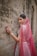A woman adorned in traditional attire stands gracefully against a textured stone wall. Her outfit features intricate embroidery and vibrant patterns. A sheer, pink veil drapes over her head and shoulders, complemented by elegant jewelry including earrings and a headpiece. Her hand is gently touching the wall, displaying decorative bangles and rings.