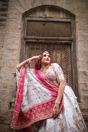 A person wears a traditional embellished outfit featuring intricate patterns in pink and white hues. The garment includes a draped fabric, ornate jewelry, and a headpiece, complementing the architectural background of an old brick wall and a wooden window with a mesh. The pose is elegant, with one arm raised.