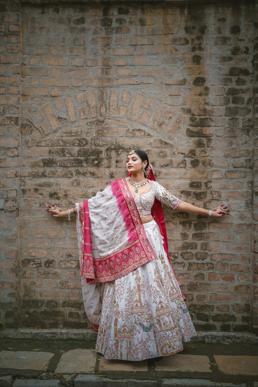 a woman in a white and pink lehenga standing in front of a brick