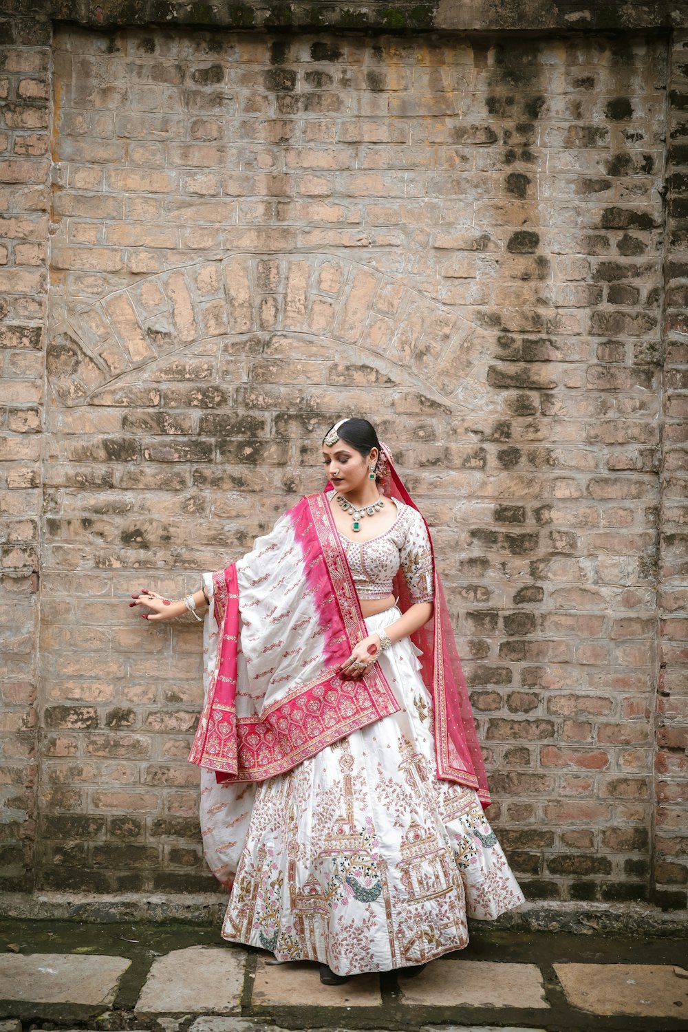 a woman in a white and pink lehenga standing in front of a brick