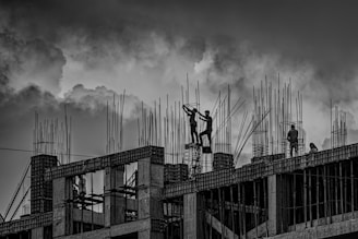 Several construction workers are on top of a building framework surrounded by numerous steel reinforcements. The sky is overcast with dramatic clouds, providing a stark and intense atmosphere to the scene.