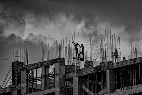 Several construction workers are on top of a building framework surrounded by numerous steel reinforcements. The sky is overcast with dramatic clouds, providing a stark and intense atmosphere to the scene.
