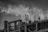 Several construction workers are on top of a building framework surrounded by numerous steel reinforcements. The sky is overcast with dramatic clouds, providing a stark and intense atmosphere to the scene.