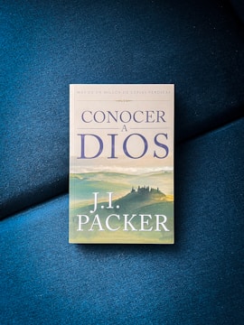 A book titled 'Conocer a Dios' by J.I. Packer is placed on a textured blue surface. The cover depicts an illustration of a castle or large building set against a scenic landscape of rolling hills with muted, earthy tones.
