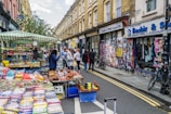 A vibrant street market with stalls filled with various goods like colorful stationery and snacks. Several people are engaged in browsing and purchasing items. The street is flanked by graffiti-covered buildings and shops. Bicycles are parked nearby, indicating a lively urban environment.