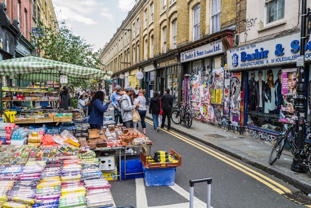 A vibrant street market with stalls filled with various goods like colorful stationery and snacks. Several people are engaged in browsing and purchasing items. The street is flanked by graffiti-covered buildings and shops. Bicycles are parked nearby, indicating a lively urban environment.