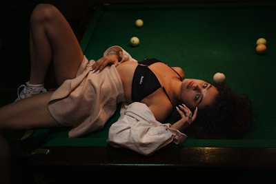 A woman is lying on a pool table, dressed in casual attire including a black top and a beige sweater. Billiard balls are scattered around the green felt surface, adding a playful yet relaxed atmosphere. The setting is dimly lit, creating a warm and intimate ambiance.