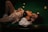A woman is lying on a pool table, dressed in casual attire including a black top and a beige sweater. Billiard balls are scattered around the green felt surface, adding a playful yet relaxed atmosphere. The setting is dimly lit, creating a warm and intimate ambiance.