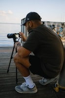 A person in casual clothing crouches on a wooden pier next to a lake, focusing intently on a camera mounted on a tripod. In the background, a bicycle is parked against a railing, and the water appears calm under a clear sky.