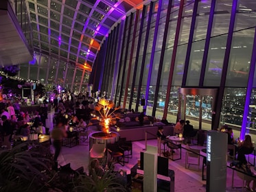 A modern indoor space with large glass windows offers a panoramic view of a cityscape at night. The interior features several groups of people seated at tables, engaging in conversation and dining. Ambient lighting in shades of purple and orange adds a warm and inviting atmosphere, with a prominent illuminated plant in the center. The bar area is bustling, contributing to a vibrant social scene.