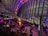 A modern indoor space with large glass windows offers a panoramic view of a cityscape at night. The interior features several groups of people seated at tables, engaging in conversation and dining. Ambient lighting in shades of purple and orange adds a warm and inviting atmosphere, with a prominent illuminated plant in the center. The bar area is bustling, contributing to a vibrant social scene.