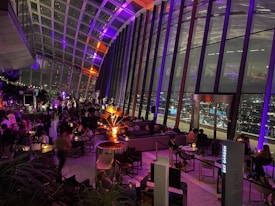 A modern indoor space with large glass windows offers a panoramic view of a cityscape at night. The interior features several groups of people seated at tables, engaging in conversation and dining. Ambient lighting in shades of purple and orange adds a warm and inviting atmosphere, with a prominent illuminated plant in the center. The bar area is bustling, contributing to a vibrant social scene.