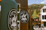 A rustic coffee stand or shop featuring a round sign with a cowboy on horseback and text 'The Coffee Cowboy, Telluride, CO'. The backdrop includes a mountainous landscape, a white house with black windows, and several people walking in casual attire.
