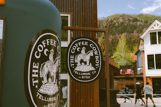 A rustic coffee stand or shop featuring a round sign with a cowboy on horseback and text 'The Coffee Cowboy, Telluride, CO'. The backdrop includes a mountainous landscape, a white house with black windows, and several people walking in casual attire.