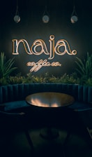 A dimly lit interior with a neon sign reading 'naja coffee co.' above a circular wooden table. The sign is surrounded by hanging pendant lights and green plants, adding a cozy and intimate vibe to the space.