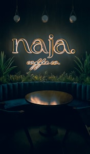 A dimly lit interior with a neon sign reading 'naja coffee co.' above a circular wooden table. The sign is surrounded by hanging pendant lights and green plants, adding a cozy and intimate vibe to the space.