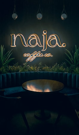 A dimly lit interior with a neon sign reading 'naja coffee co.' above a circular wooden table. The sign is surrounded by hanging pendant lights and green plants, adding a cozy and intimate vibe to the space.