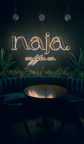 A dimly lit interior with a neon sign reading 'naja coffee co.' above a circular wooden table. The sign is surrounded by hanging pendant lights and green plants, adding a cozy and intimate vibe to the space.
