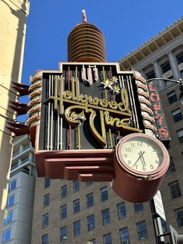 A vintage neon sign with the words 'Hollywood and Vine' in bright yellow cursive, accompanied by a clock showing the time just after 3 o'clock. The sign features a classic retro design against the backdrop of tall urban buildings and a clear blue sky.