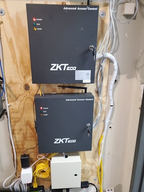 A wall-mounted setup featuring two black electronic control boxes labeled 'Advanced Access Control' by ZKTeco. Each box has indicator lights for power, run, and communication. The boxes are mounted on a wooden panel, surrounded by a variety of cables including yellow and white ones. The smaller box is mounted below the larger one and both have key locks.