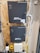 A wall-mounted setup featuring two black electronic control boxes labeled 'Advanced Access Control' by ZKTeco. Each box has indicator lights for power, run, and communication. The boxes are mounted on a wooden panel, surrounded by a variety of cables including yellow and white ones. The smaller box is mounted below the larger one and both have key locks.