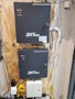 A wall-mounted setup featuring two black electronic control boxes labeled 'Advanced Access Control' by ZKTeco. Each box has indicator lights for power, run, and communication. The boxes are mounted on a wooden panel, surrounded by a variety of cables including yellow and white ones. The smaller box is mounted below the larger one and both have key locks.