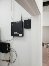 A security and access control system is mounted on a wall with visible wiring and electronic equipment. The ZKTeco brand is prominently displayed on the black access control box. Additional equipment is present, including a network switch with connected cables. There are paint supplies and a pink protective floor covering in the adjacent room.