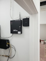A security and access control system is mounted on a wall with visible wiring and electronic equipment. The ZKTeco brand is prominently displayed on the black access control box. Additional equipment is present, including a network switch with connected cables. There are paint supplies and a pink protective floor covering in the adjacent room.