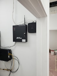 A security and access control system is mounted on a wall with visible wiring and electronic equipment. The ZKTeco brand is prominently displayed on the black access control box. Additional equipment is present, including a network switch with connected cables. There are paint supplies and a pink protective floor covering in the adjacent room.