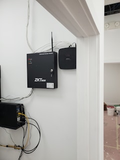 A security and access control system is mounted on a wall with visible wiring and electronic equipment. The ZKTeco brand is prominently displayed on the black access control box. Additional equipment is present, including a network switch with connected cables. There are paint supplies and a pink protective floor covering in the adjacent room.