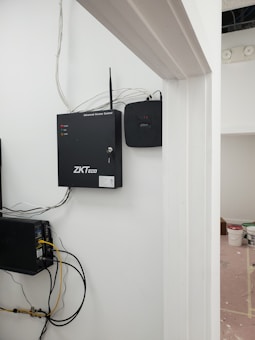 A security and access control system is mounted on a wall with visible wiring and electronic equipment. The ZKTeco brand is prominently displayed on the black access control box. Additional equipment is present, including a network switch with connected cables. There are paint supplies and a pink protective floor covering in the adjacent room.