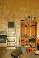 A room with a worn, textured wall and scattered tools. A metal ladder leans against the wall next to a wooden shelving unit containing various items like a drill, packing materials, and paint cans. The wooden floor is covered with more tools and a toolbox, suggesting ongoing renovation or maintenance work.