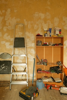 A room with a worn, textured wall and scattered tools. A metal ladder leans against the wall next to a wooden shelving unit containing various items like a drill, packing materials, and paint cans. The wooden floor is covered with more tools and a toolbox, suggesting ongoing renovation or maintenance work.