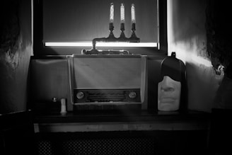 A vintage radio with a dial and knobs sits on a table against a wall. Above the radio, there are three exposed lightbulbs mounted with pipes. To the right of the radio, a white mask with a contemplative facial expression is placed. The scene is dimly lit, and the image is black and white.