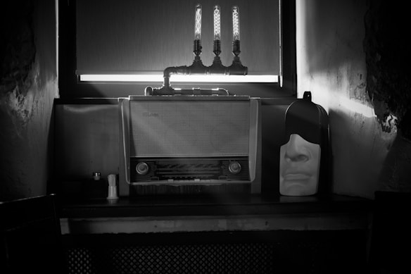 A vintage radio with a dial and knobs sits on a table against a wall. Above the radio, there are three exposed lightbulbs mounted with pipes. To the right of the radio, a white mask with a contemplative facial expression is placed. The scene is dimly lit, and the image is black and white.