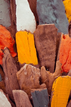 Several pieces of painted wooden planks arranged vertically, showcasing a variety of colors including orange, yellow, gray, white, and natural wood tones. The surfaces appear textured and weathered, adding a rustic and artistic feel to the scene.