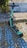 A teal electric scooter is parked on a cobblestone walkway with patches of blue-painted stones. Grass is growing between the stones, and there is a concrete wall in the background partially covered by vegetation.