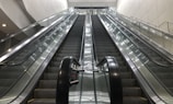 A set of parallel escalators lead upward, flanked by shiny glass railings and metallic side panels. The architecture appears modern, with clean lines and a bright color palette. Overhead lighting provides even illumination, enhancing the sleek and functional design.