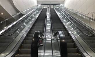 A set of parallel escalators lead upward, flanked by shiny glass railings and metallic side panels. The architecture appears modern, with clean lines and a bright color palette. Overhead lighting provides even illumination, enhancing the sleek and functional design.