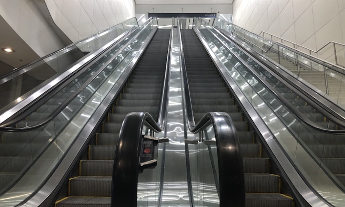 A set of parallel escalators lead upward, flanked by shiny glass railings and metallic side panels. The architecture appears modern, with clean lines and a bright color palette. Overhead lighting provides even illumination, enhancing the sleek and functional design.