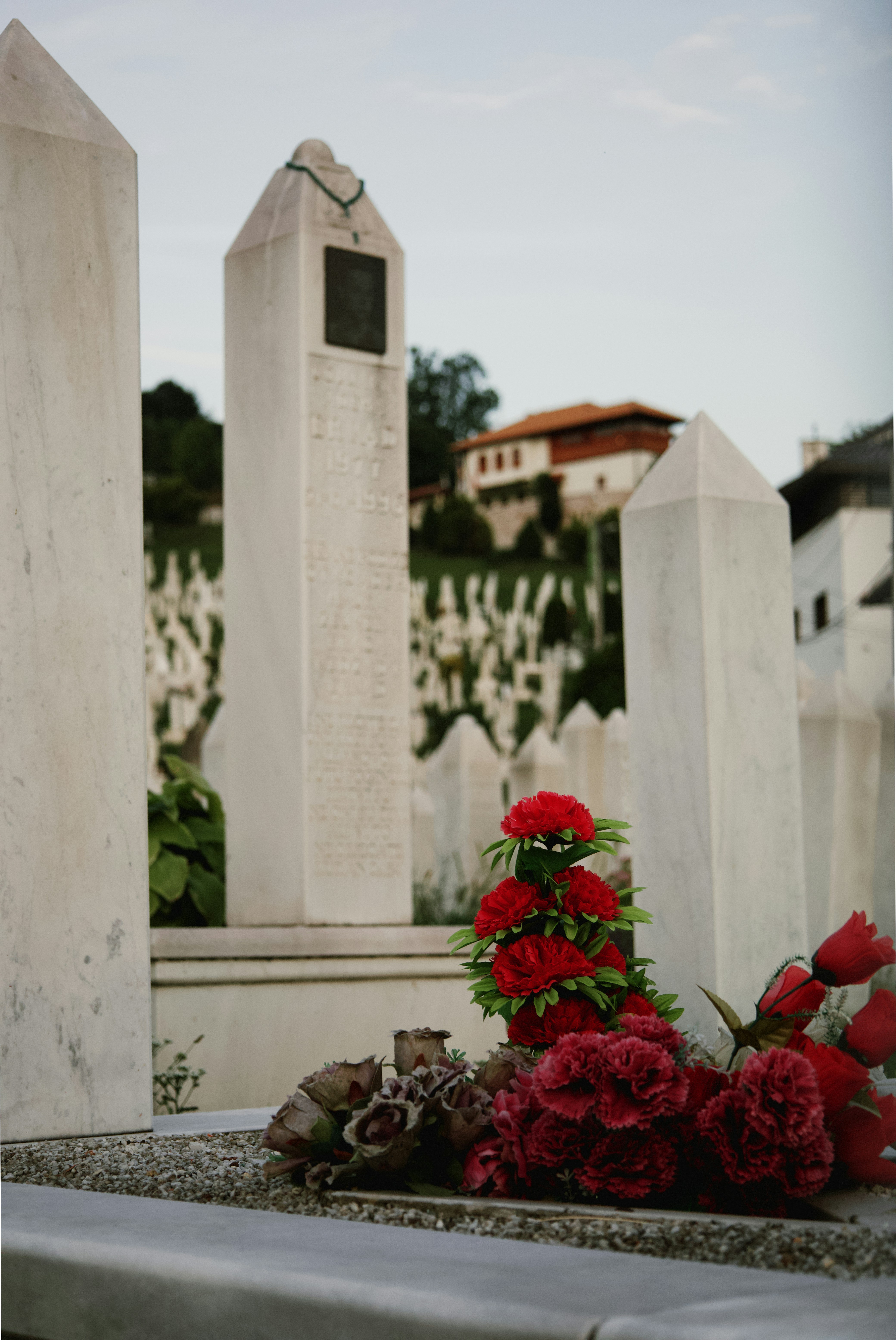 A cemetery with a focus on a single grave adorned with red flowers, surrounded by white marble tombstones featuring inscriptions. The background includes a hillside filled with similar tombstones and a few buildings.