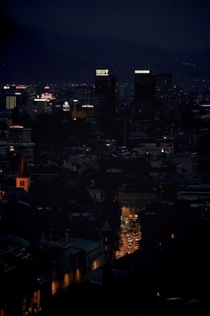 A nighttime cityscape featuring illuminated skyscrapers with prominent logos on top, surrounded by a mixture of tall buildings and smaller structures. Streets are visible with lines of cars, and the city is set against a backdrop of dark hills.