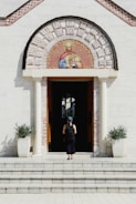 A person wearing a blue cap and backpack stands in front of a church entrance, which features two columns and an ornate semicircular mosaic depicting religious figures above the doorway. Potted plants flank the entrance on either side.