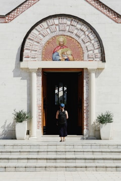 A person wearing a blue cap and backpack stands in front of a church entrance, which features two columns and an ornate semicircular mosaic depicting religious figures above the doorway. Potted plants flank the entrance on either side.