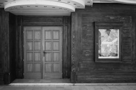 A black and white image of a vintage wooden door with intricate paneling, set within a wooden exterior wall. There is a reflective glass frame to the right of the door, showing an abstract reflection of outdoor elements. The ceiling above the door exhibits circular recessed lighting.